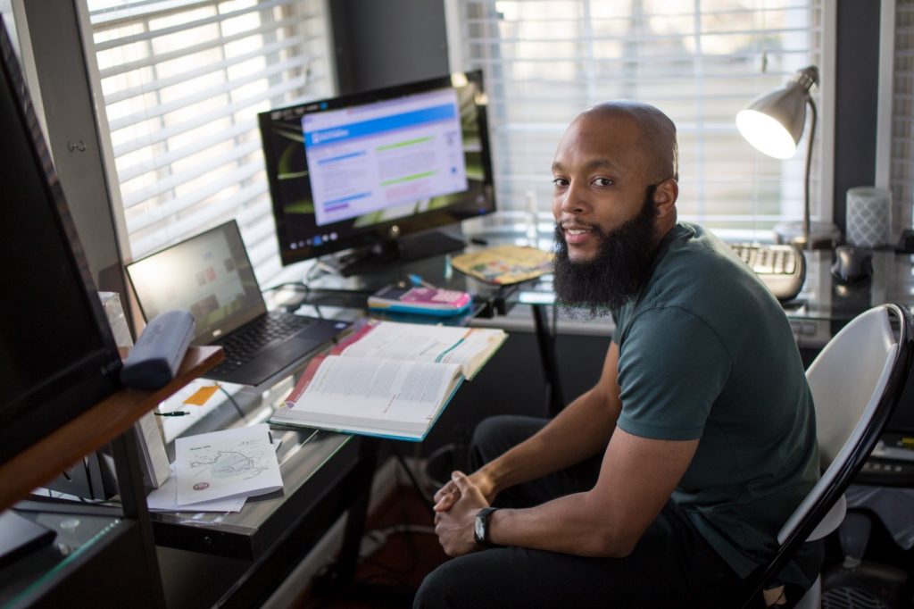 A student sits at a desk with laptop, open textbook, and extra monitor