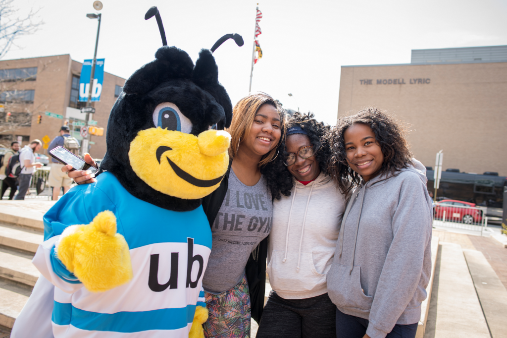 Three students pose with the UBalt mascot in an outdoor plaza